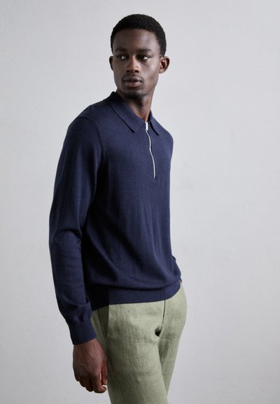 Navyblauer Strickpullover mit einem Reißverschlussdetail und spitzem Kragen, kombiniert mit hellgrünen Leinenhosen. Glatte Textur und taillierte Passform.