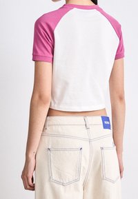 Camiseta de algodón con cuerpo blanco y mangas raglán rosas. Presenta un cuello redondo y una textura suave. Se lleva con pantalones de cintura alta beige.