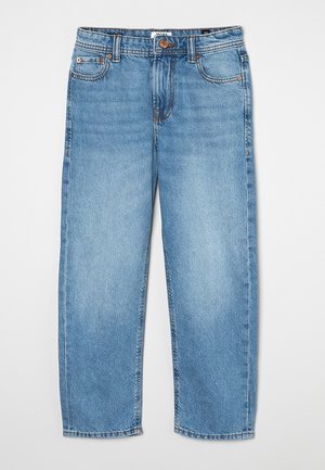 Bas des jambes en jean bleu clair portant des baskets crème avec des rayures vertes, un détail rouge au talon et des semelles en caoutchouc sur fond blanc.
