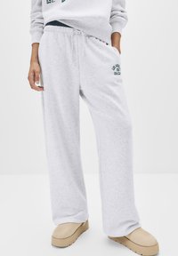 Helle graue Jogginghose aus weichem Material, ausgestattet mit einem elastischen Bund mit Kordelzug und grünem gesticktem Logo-Detail an der Seite.