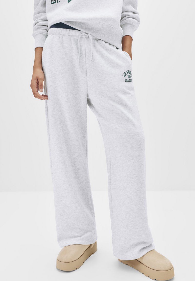 Helle graue Jogginghose aus weichem Material, ausgestattet mit einem elastischen Bund mit Kordelzug und grünem gesticktem Logo-Detail an der Seite.