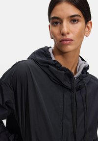 Czarna kurtka z kapturem o lekkim, matowym wykończeniu. Posiada szary wewnętrzny podszewkę i sznurki w kapturze, z luźnym krojem.