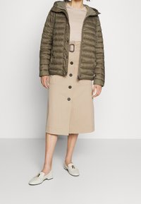 Olivově zelená péřová bunda s horizontálním prošíváním, béžová sukně na knoflíky a světlé hnědé loafers se zlatým akcentem.