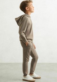 Barn iført en khaki hættetrøje og matchende joggers med sidelæns striber. Bærer hvide sneakers og står på en lys overflade.