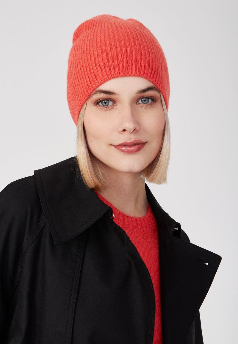 Berretto a coste rosso realizzato in materiale morbido, indossato con un cappotto nero e un maglione rosso brillante, evidenziando la texture e il contrasto dei colori.