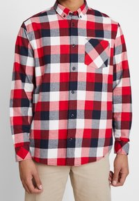 Camisa de franela con cuadros en rojo, azul marino, gris y blanco, con cuello abotonado, bolsillo frontal y mangas largas, que presenta un dobladillo curvado y botones.