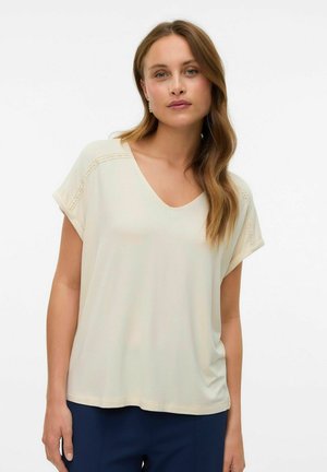Top met V-hals en korte mouwen in lichtbeige stof met een zachte textuur en gestikte details langs de schouders. Gecombineerd met marineblauwe broek.