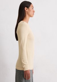 Beige longsleeve top in een aansluitend ontwerp. Gladde textuur met geribbelde details langs de mouwen en de halslijn. Gecombineerd met een grijze stoffen broek.