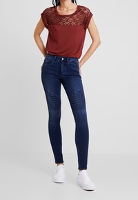Top burdeos con detalles de encaje y mangas cortas, combinado con jeans skinny azul oscuro con costuras y paneles texturizados en las rodillas.