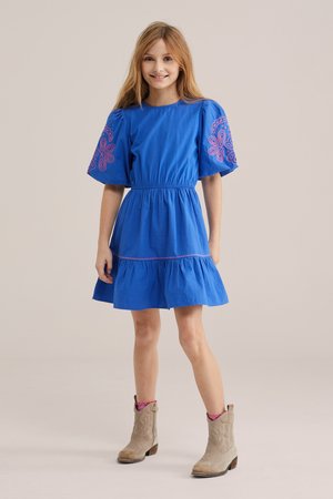 Ragazza giovane che sorride, indossa un abito blu con maniche a sbuffo e ricami, abbinato a stivaletti beige e calzini rosa.