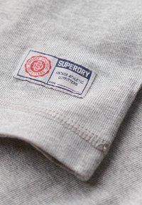 Grau-gestreifter Baumwoll-T-Shirt-Stoff mit strukturierter Oberfläche, versehen mit einem rechteckigen gestickten Etikett in Blau und Rot, das "Superdry Vintage Athletic Outfitters" liest.