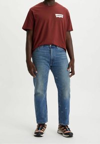 Camiseta de manga corta color marrón con el logo blanco de "Levi's", combinada con jeans rotos de color azul claro y zapatillas deportivas oscuras con detalles en naranja.