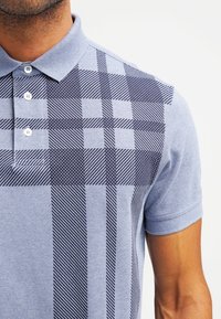 Polo en bleu clair avec motif tartan bleu foncé ; manches courtes, col boutonné, tissu texturé et motifs linéaires distincts.