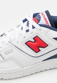 Vit sneaker med marinblå och röda accenter, läderöverdel, perforerad detalj och gummisula; har "550" och varumärkeslogotyp på sidan.