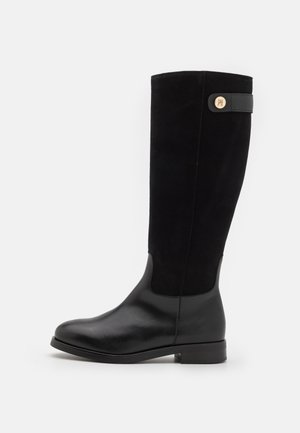 Botte noire pour femme mi-mollet avec pied en cuir lisse, tige en daim, talon bas, et bride décorative avec un emblème rond doré près du haut.