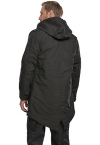 La chaqueta parka negra con capucha presenta un dobladillo trasero largo, tejido texturizado, costuras visibles y detalles de botones en la parte posterior y en las mangas.