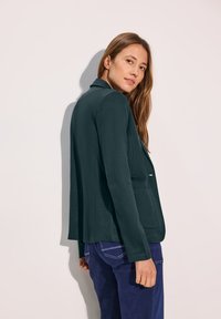Blazer verde oscuro de tejido suave, con cuello de muesca, cierre de un solo botón y dos bolsillos frontales.