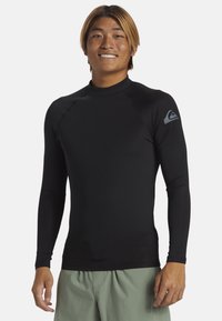 Rash guard negro de manga larga, hecho de material elástico, con un cuello alto y un acento de logo gris en el hombro izquierdo.