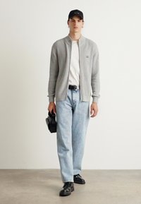 Cardigan grigio a coste, t-shirt bianca, jeans azzurri chiari, scarpe nere, in mano una borsa nera, indossa un cappellino nero. Design semplice, abbigliamento casual.