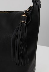Sac à main en cuir noir avec un long détail de pompon fixé par des anneaux dorés près de la fermeture éclair supérieure.