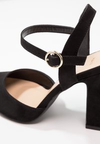 Černá semišová lodička na vysokém podpatku s širokým podpatkem, s řemínkem kolem kotníku a zlatou kulatou sponou, a kulatou špičkou. Hladká vnitřní podšívka.