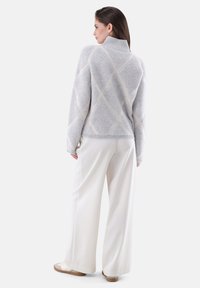 Grauer Strickpullover mit hohem Kragen, mit einem diamantförmigen Muster in hellbeige. Kombiniert mit weißen weit geschnittenen Hosen und hellen Schuhen.