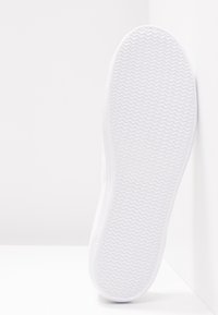 Baskets blanches avec une semelle en caoutchouc texturée et un motif en zigzag, un matériau supérieur lisse et un design bas. Vue de côté sur fond clair.