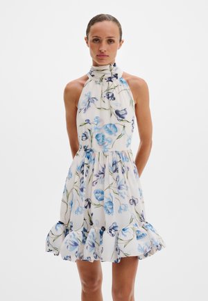 Nő ujjatlan fehér ruhában, kék virágmintával és fodros szegéllyel, fehér háttér előtt áll, kezei a háta mögött.