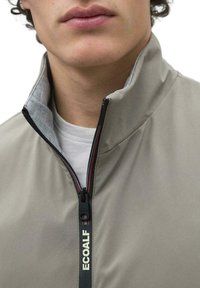 Chaqueta gris claro con cremallera y un alto cuello. Presenta una cremallera negra con acentos rojos y la marca ECOALF, llevada sobre una camisa blanca.