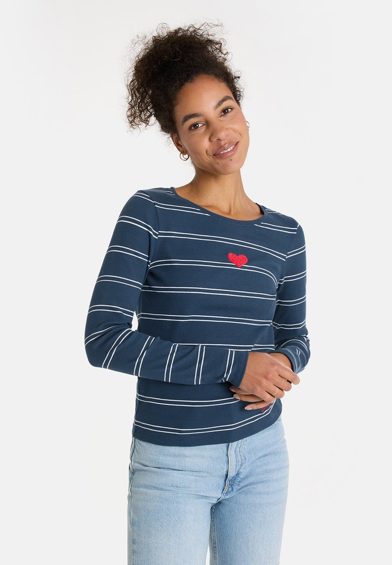Frau mit lockigem Haar, die ein navyblaues und weiß gestreiftes Langarmshirt mit einem roten Herzen auf der Brust und hellblauen Jeans trägt und sanft lächelt.