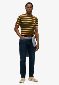 Grøn stribet t-shirt med lodrette orange, gule og hvide striber, parret med mørke jeans og lyse sneakers. T-shirten har logodetaljer.