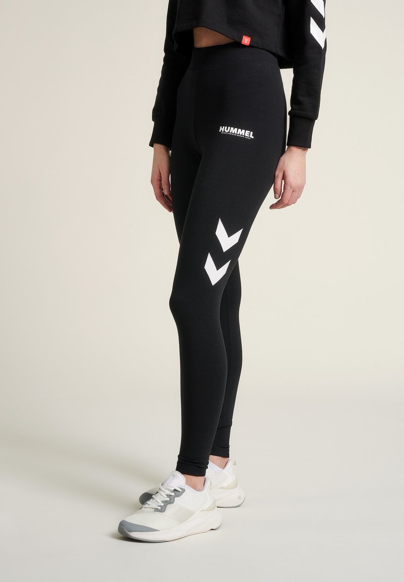 Leggings noirs à taille haute, arborant le logo blanc Hummel et des motifs en chevrons sur les côtés. Associés à des baskets blanches légères.