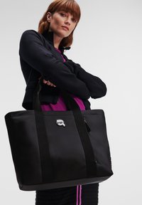 Zwarte schoudertas van textuurstof, met dubbele handgrepen en een klein logo-accent. Beschikt over een ruime binnenkant en een ritsvak.