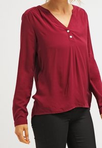 Blusa de manga larga color burdeos con cuello en V, tres botones decorativos en el cuello y un ajuste ligeramente holgado. Textura suave, diseño sencillo.