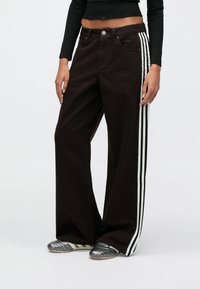 Καφέ τζιν παντελόνι με φαρδιά μπάσκα και λευκές πλευρικές ρίγες, κατασκευασμένο από βαμβάκι. Διαθέτει πέντε τσέπες και κλείσιμο με κουμπί. Φοριέται με μεταλλικά παπούτσια.