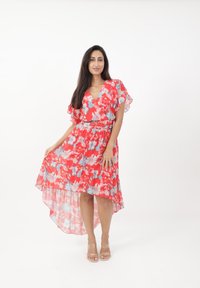 Robe florale rouge avec des manches courtes en volants, décolleté en V, taille froncée et ourlet asymétrique. Assortie à des sandales transparentes à lanières.