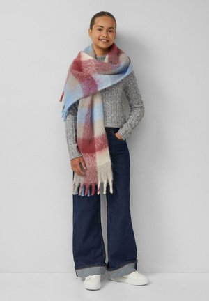 Écharpe à motif écossais en mélange de laine douce, mettant en avant des couleurs comme le rouge, le bleu et le beige. Les bords à franges ajoutent de la texture. Le modèle porte un pull gris et un jean.
