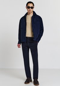 Giacca in suede blu navy con due tasche al petto sopra un maglione beige in maglia, abbinata a pantaloni in corduroy blu navy scuro e scarpe stringate marroni.