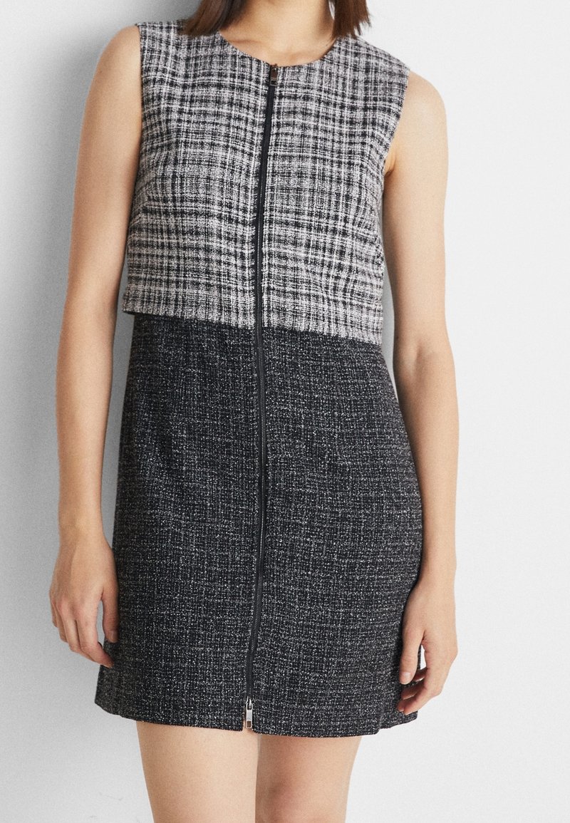 Robe sans manches longueur genou avec fermeture éclair frontale, comportant la moitié supérieure à carreaux gris et noir et la moitié inférieure noire unie texturée.