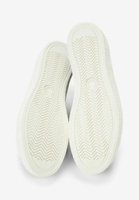 Suelas de zapatillas blancas con goma texturizada para agarre, que presentan un patrón en zigzag y un logo circular en el centro.