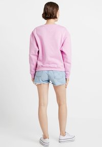 Rosa sweatshirt med avslappnad passform, ribbad mudd och nederkant, tillsammans med ljusblå slitna denimshorts. Modellen bär vita sneakers.