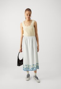 Strikket beige topp med bølgekant i halsen, kombinert med et langt hvitt skjørt med blå blomstertrykk og en svart håndveske. Grå sneakers på føttene.