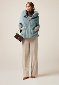Cappotto in pelliccia sintetica blu con cappuccio, chiusura con zip e tasche laterali. Indossato sopra un dolcevita grigio, abbinato a pantaloni a gamba larga beige e una borsa marrone.