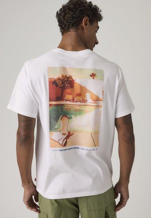 Homme aux cheveux bouclés portant un t-shirt blanc avec une scène au bord de la piscine illustrant des chaises, un arbre et les jambes d'une personne étendues à l'arrière.