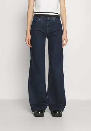 Osoba v tmavě modrých širokých džínách, bílém krátkém rukávu s černým lemováním a černých platformových botách na jednoduchém pozadí.