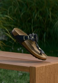 Μαύρο σανδάλι με λουλουδένιες λεπτομέρειες, πάτο από φελλό, ρυθμιζόμενο λουράκι, υφή στη σόλα, τοποθετημένο πάνω σε θολό φυσικό φόντο.