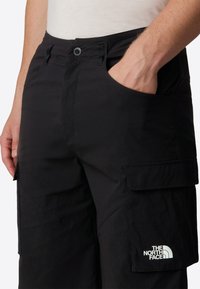 Close-up of a person wearing black cargo shorts with front pockets, one hand inside a pocket, and "The North Face" logo on the leg pocket.