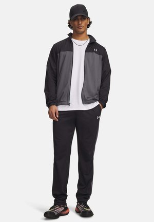 Man met een zwarte pet, een grijs-zwart jack met rits, een wit T-shirt, zwarte broek en zwarte sneakers, staand met één hand in zijn zak.