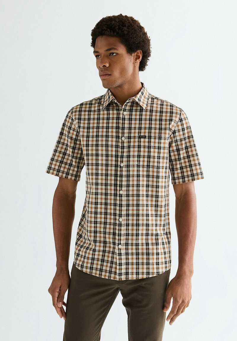 Uomo che indossa una camicia beige, nera e bianca a quadri con maniche corte e pantaloni marrone scuro, in piedi contro uno sfondo chiaro e semplice.