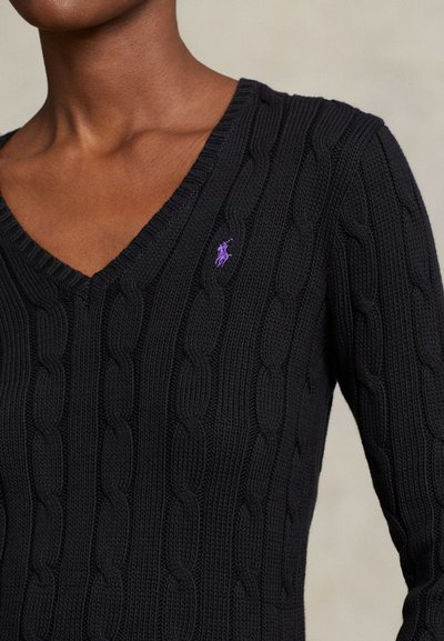 Približen posnetek osebe, ki nosi črni pulover s kabelsko pletenim V-izrezom in majhnim vijoličnim vezenim logotipom na levi strani prsnega koša.